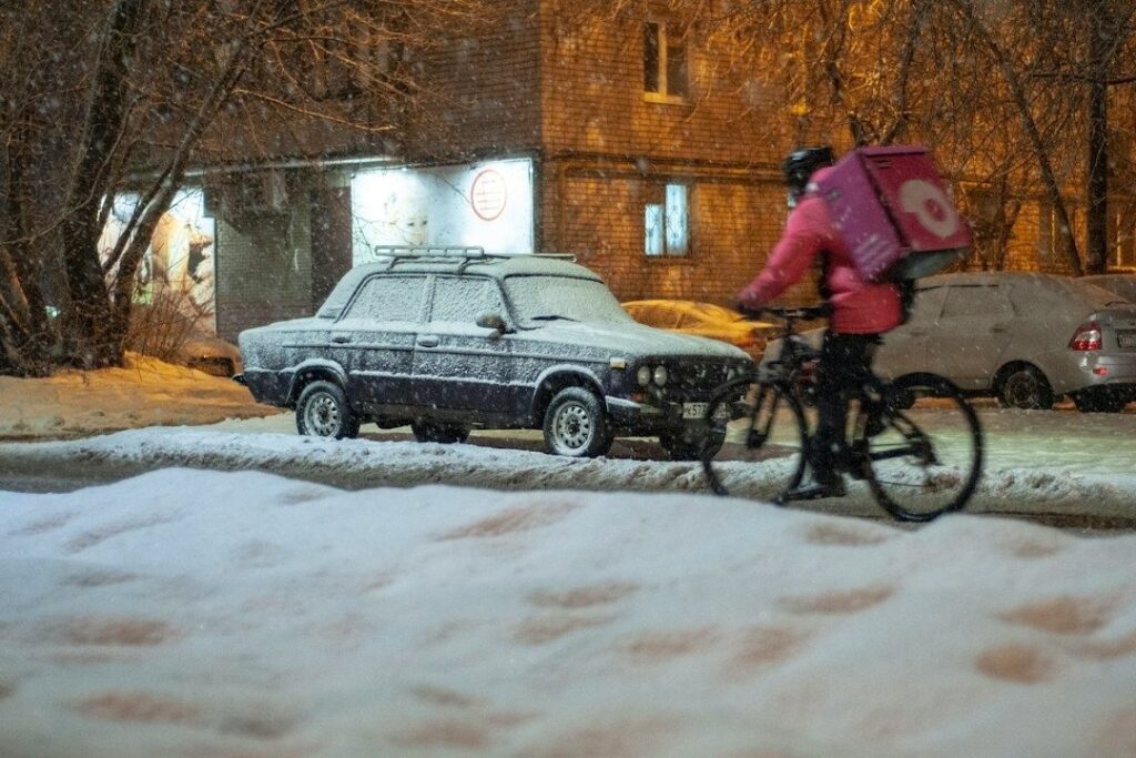 Cykelbud med rosa leveransväska cyklar i snö förbi en frostig bil