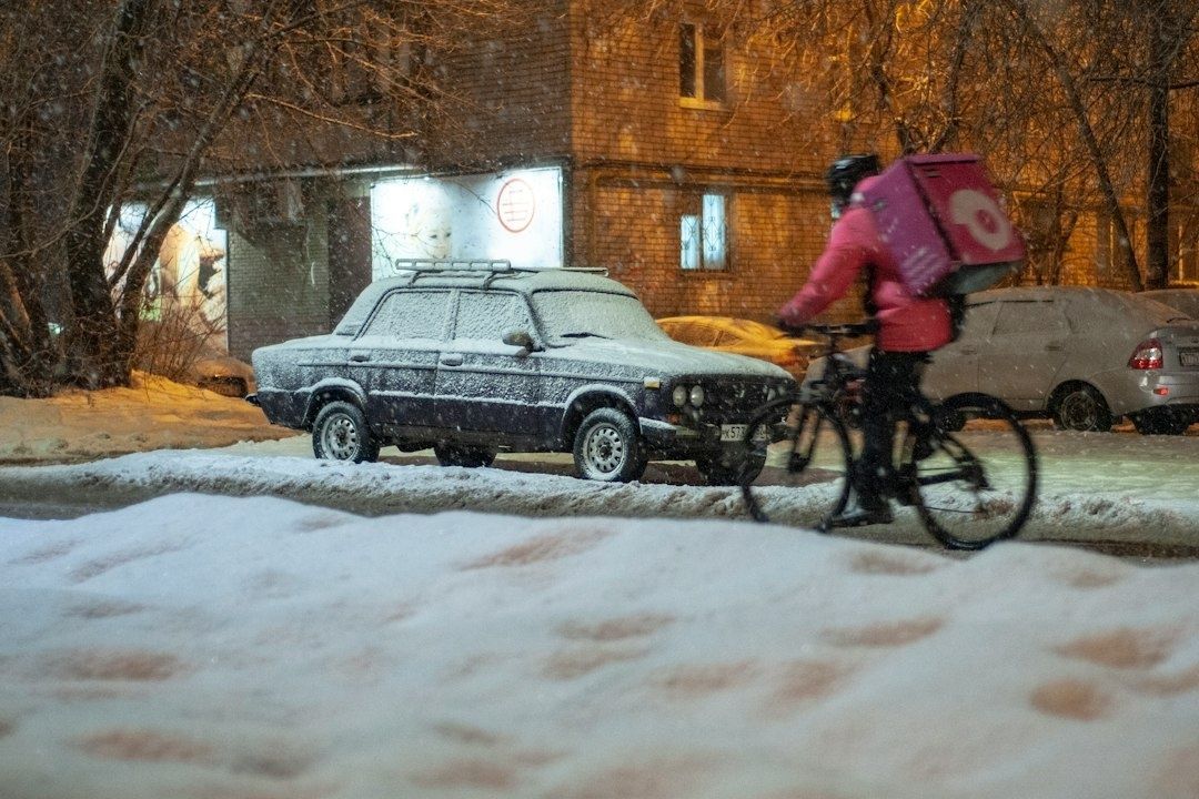 Cykelbud med rosa leveransväska cyklar i snö förbi en frostig bil